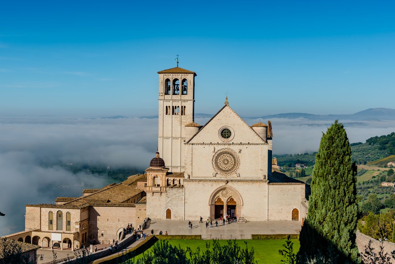 Basilica di San Francesco in Assisi, Italy, with misty hill views and vibrant blue sky.